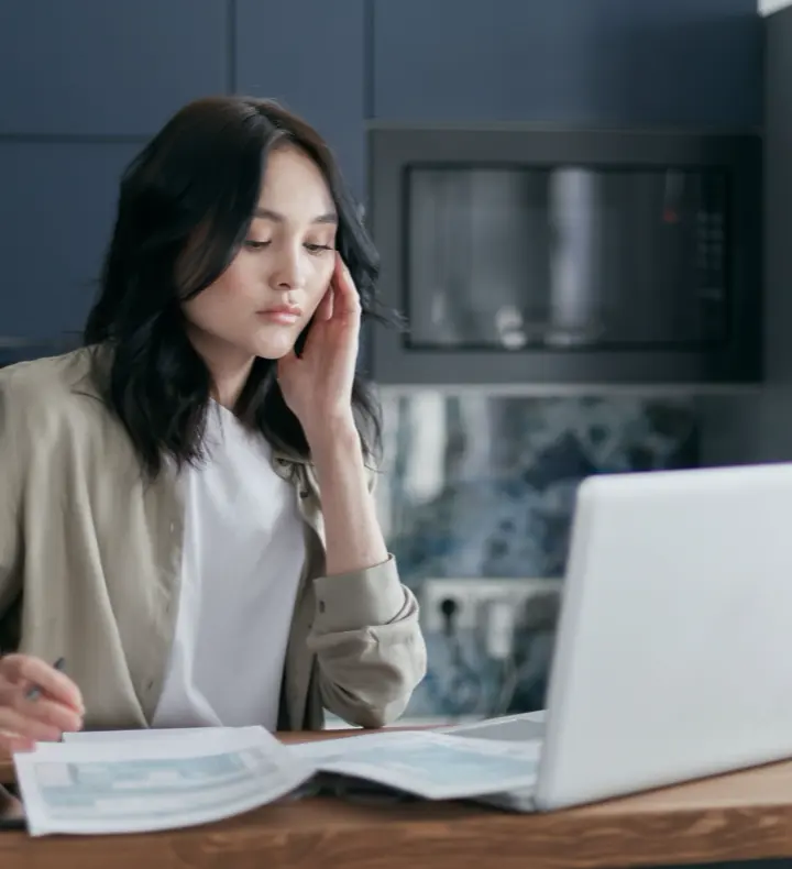A woman at a kitchen table comparing paperwork against her laptop.