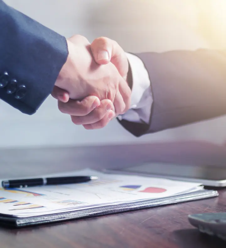 Two people shaking hands across a desk of financial charts.